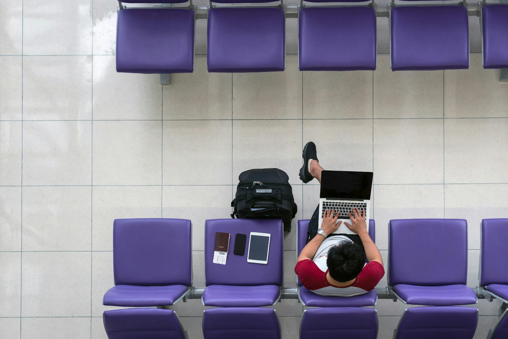 An aerial shot of a male traveller wearing shorts and tshirt working on his laptop in an airport departure lounge. His tablet, phone and passport are on a purple seat next to him and his bag is on the ground.