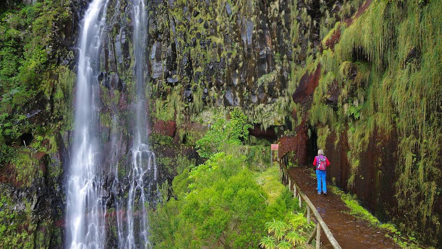 A woman stands by a levada, an open irrigation channel specific to Madeira, looking at a waterfall amid dense greenery and leafy scenery. She wears a colourful backpack, bright blue trousers and a purple gilet over a red long-sleeved top.