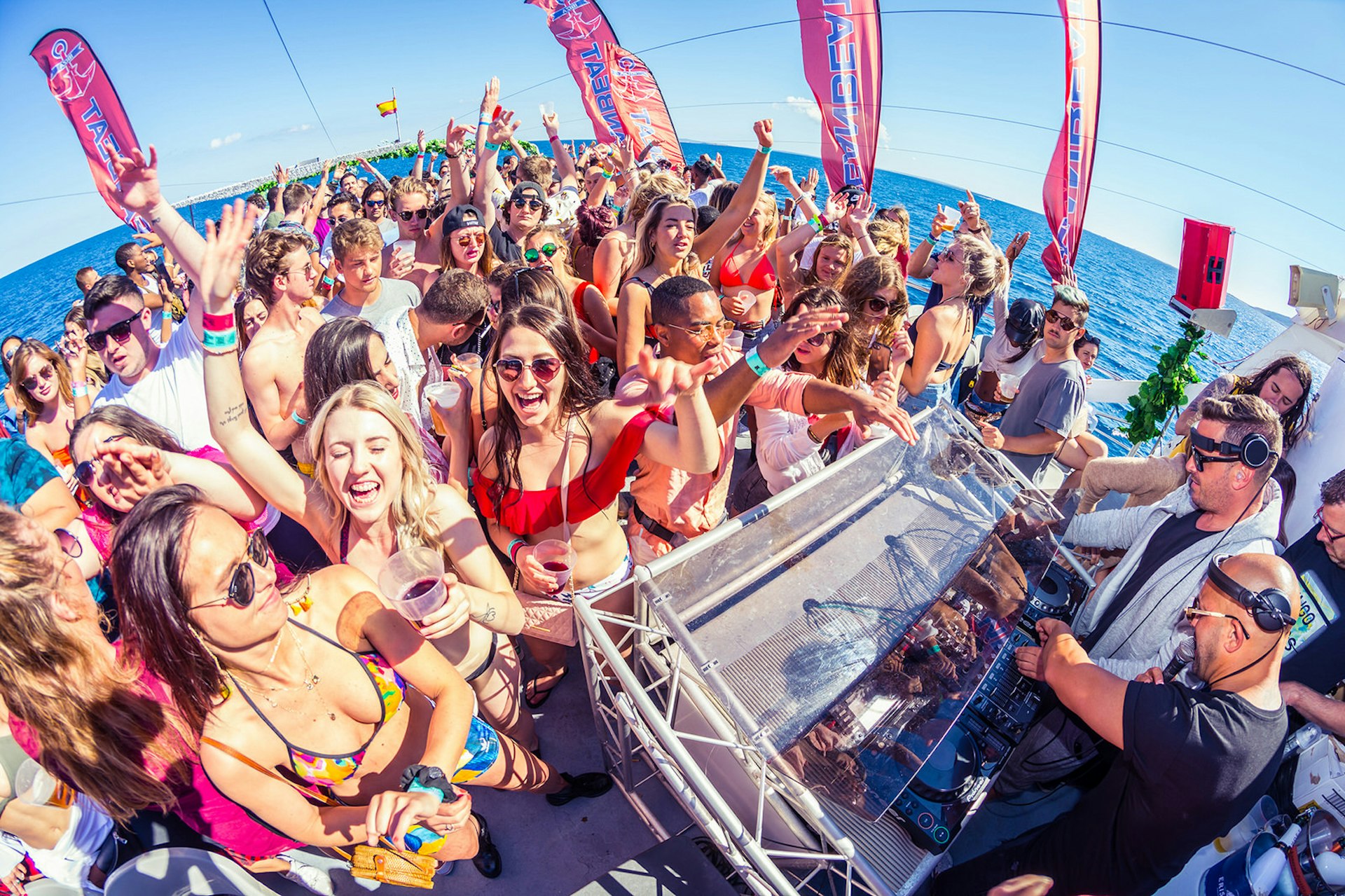 A large crowd on the deck of a boat gather around a DJ deck with two men wearing headphones. The crowd are wearing swimwear and have their hands in the air, there are large red flags saying 'Ocenbeat' around the sides of the boat