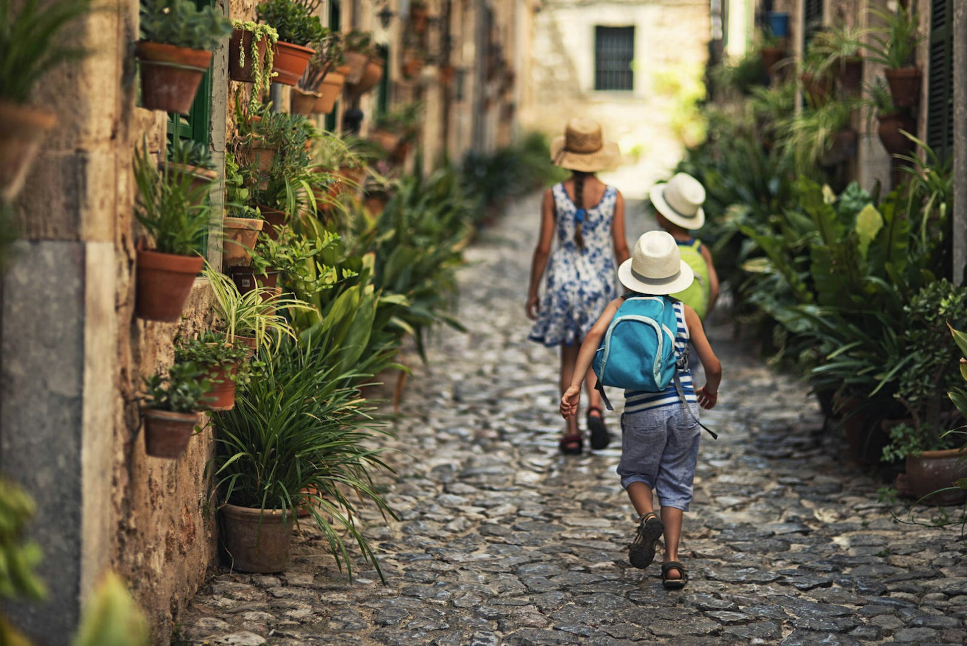 Three kids walking away from the camera in a cobble-stone alley lined with potted plants; the kids are all wearing sunhats and two have backpacks