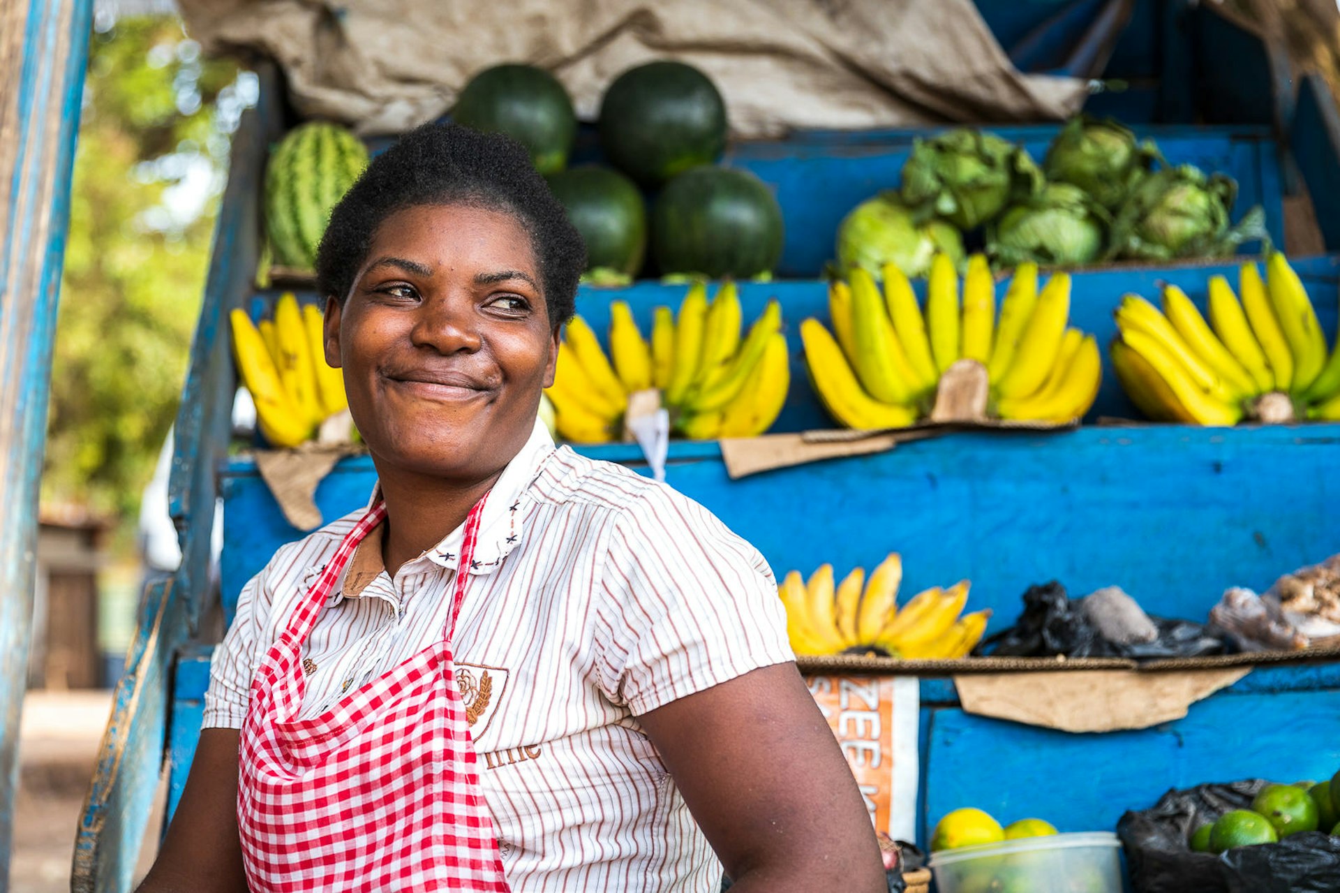 A Ugandan market trader grins, with rows of bananas stacked behind her