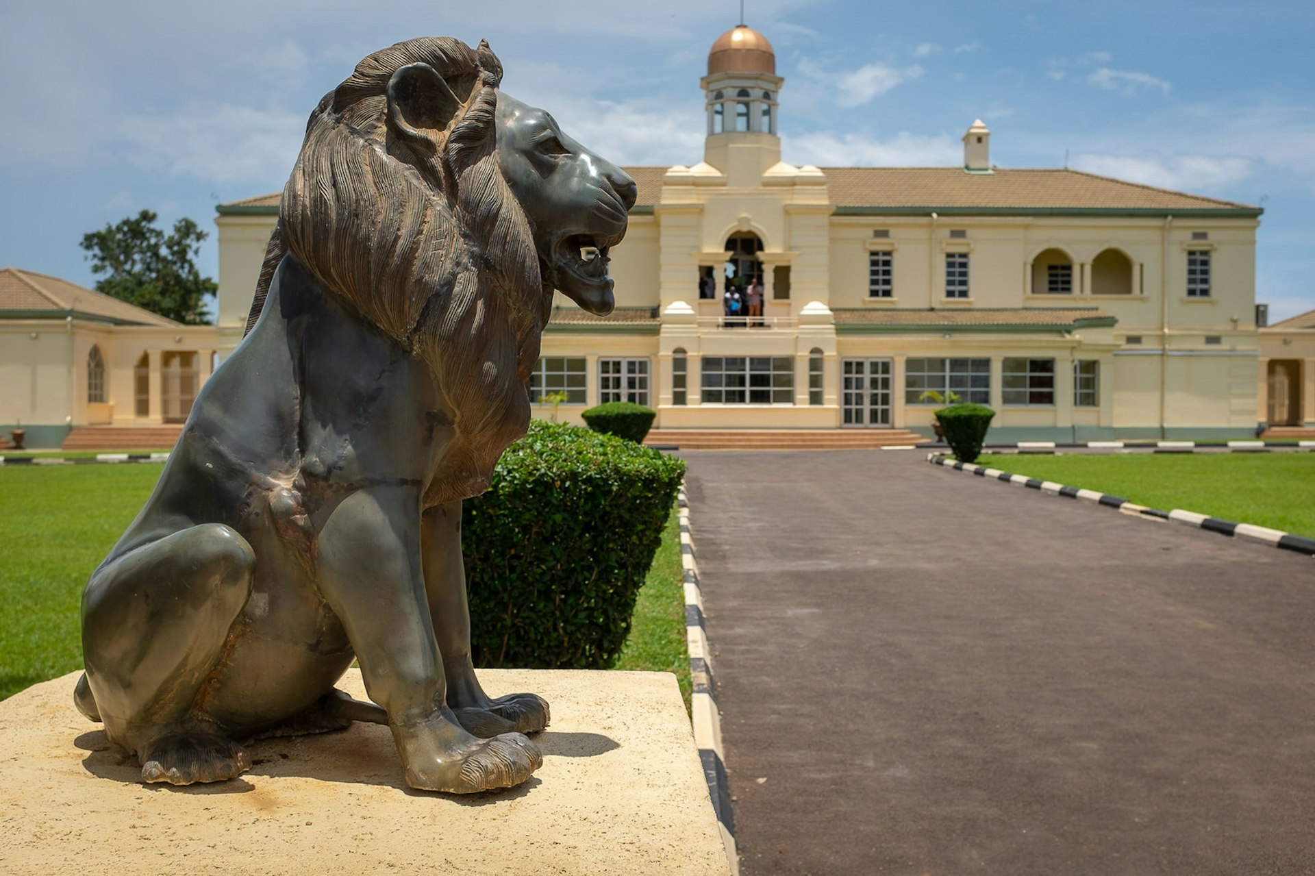 A large stone lion stands next to a paved path leading to Mengo Palace in the distance; the cream-coloured building is grand, with a gold-topped tower