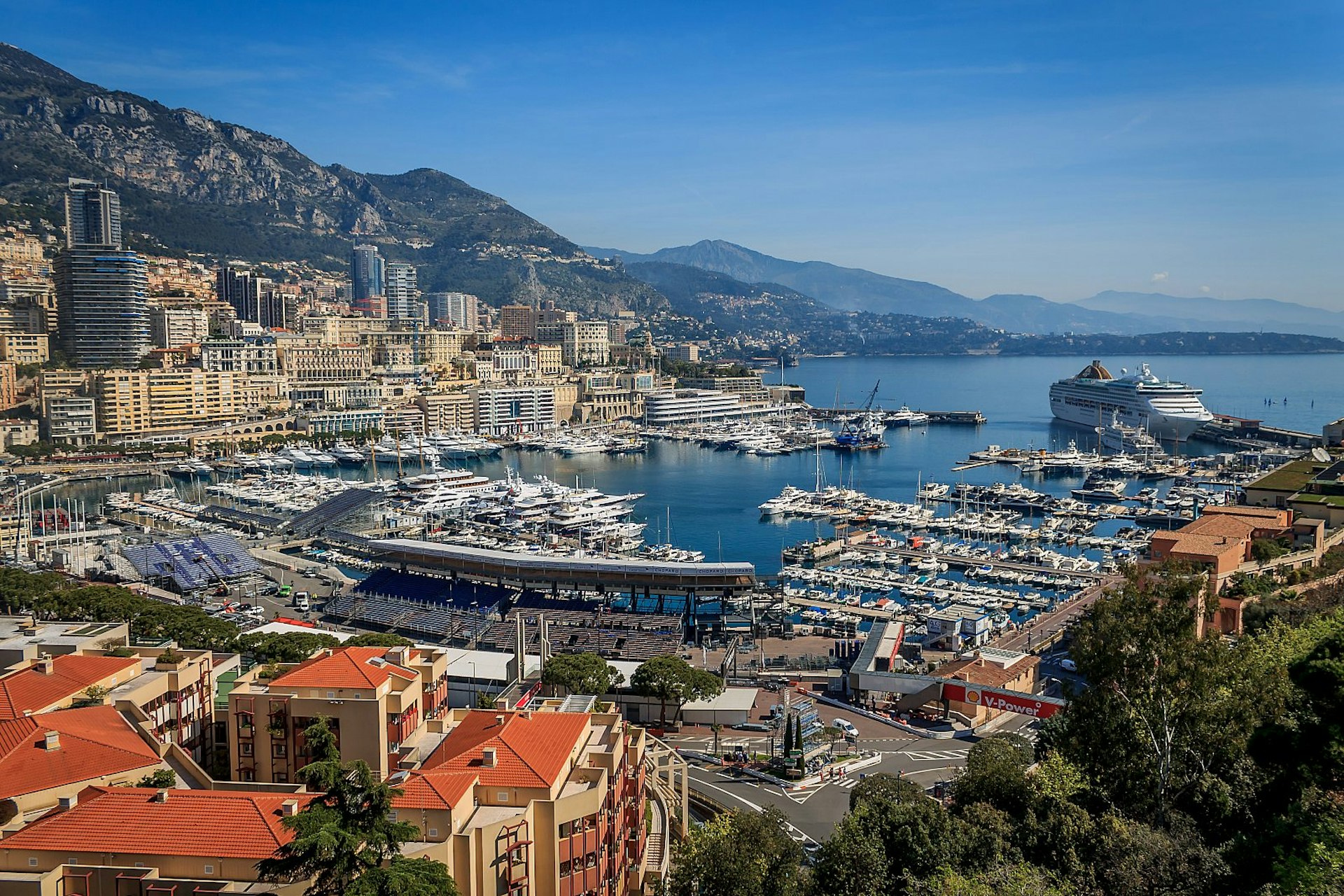 A panoramic view of the yacht-filled harbour and modern skyline of Monaco; the coastline is hugged by mountains stretching into the distance. 
