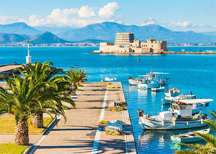 A paved jetty lined with palm trees, with boats next to it in the harbour; beyond is a fortress on an islet, with mountains on the coastline further away still.