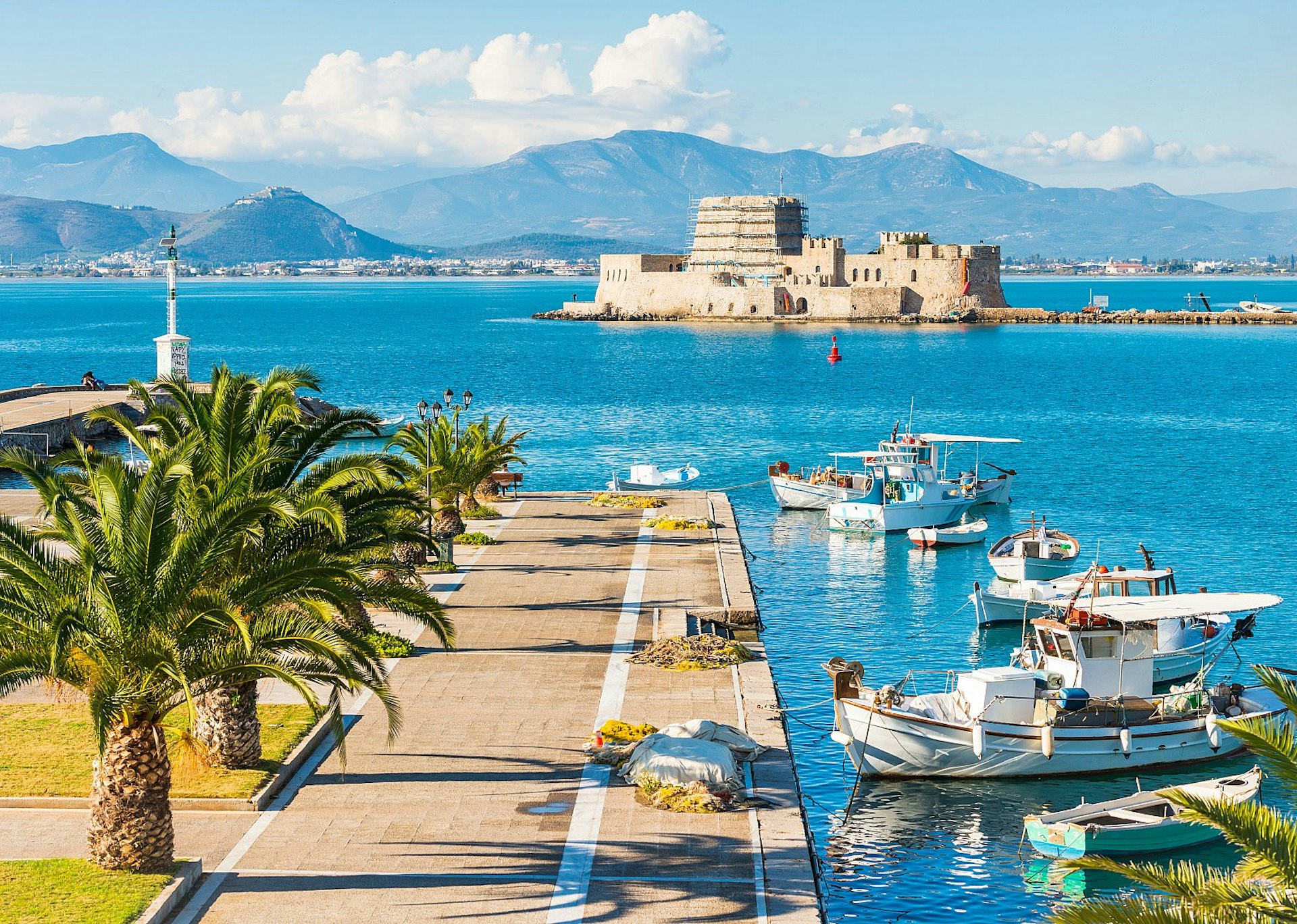 A paved jetty lined with palm trees, with boats next to it in the harbour; beyond is a fortress on an islet, with mountains on the coastline further away still.