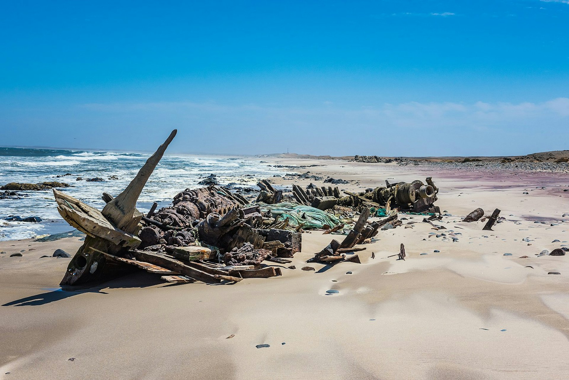 A wooden shipwreck on a huge empty sand beach along Namibia's Skeleton Coast, while a powerful-looking Atlantic Ocean laps the shore. 