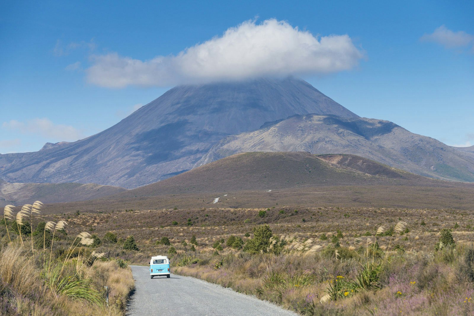 Campervan on road in Tongariro National Park, with volcanic peak (with its rocky summit cloaked in a petite cloud, in background