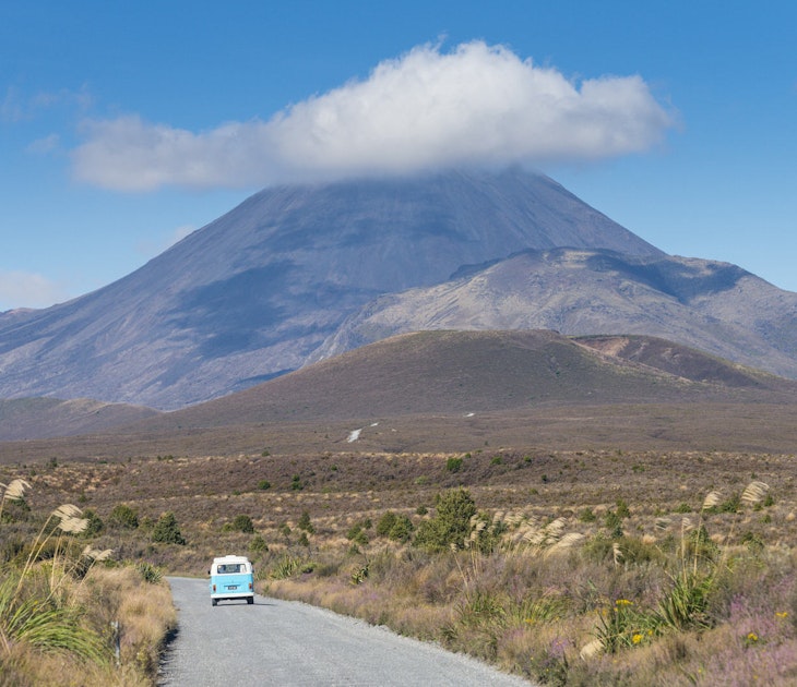 Campervan on road in Tongariro National Park, with volcanic peak (with its rocky summit cloaked in a petite cloud, in background