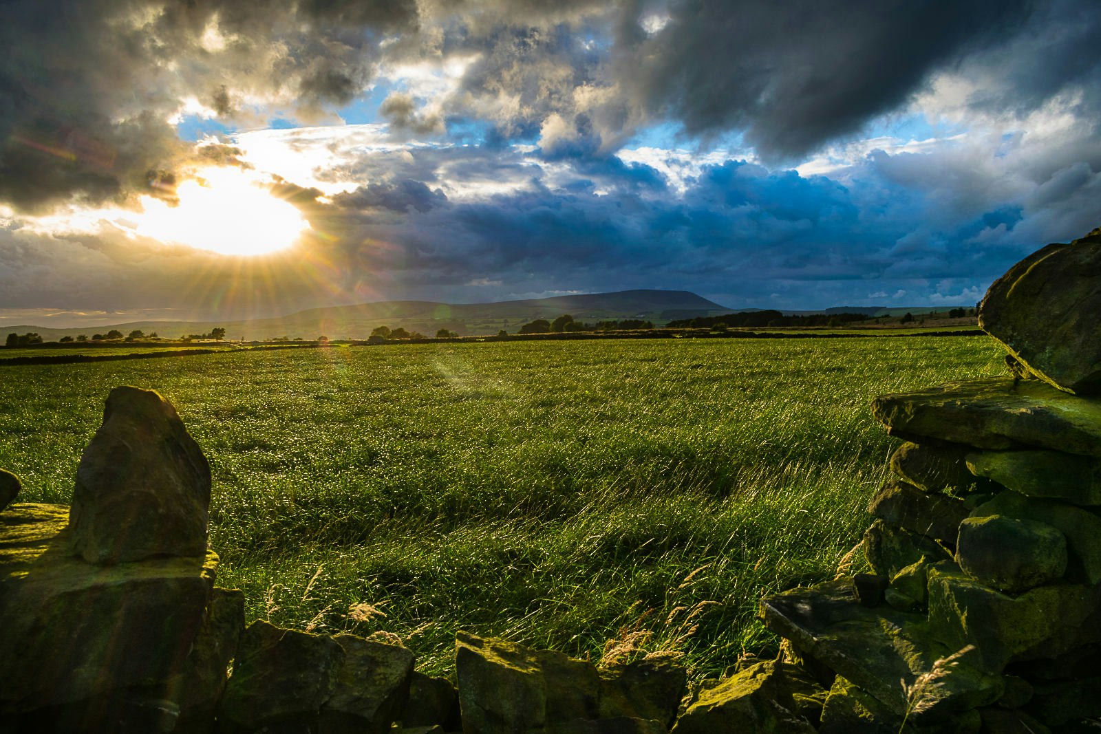A stone wall at the foreground of the image framing grass and the sun peeking out from behind cloud cover with a hill in shadow in the background.