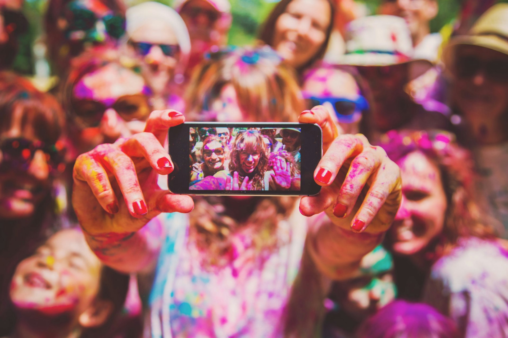 A group of friends covered in multi-coloured powder are out of focus while one of them holds a phone to take a selfie. The smaller image of them is sharper and you can see them wave to the camera