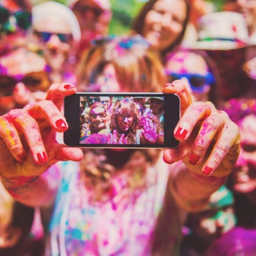 A group of friends covered in multi-coloured powder are out of focus while one of them holds a phone to take a selfie. The smaller image of them is sharper and you can see them wave to the camera