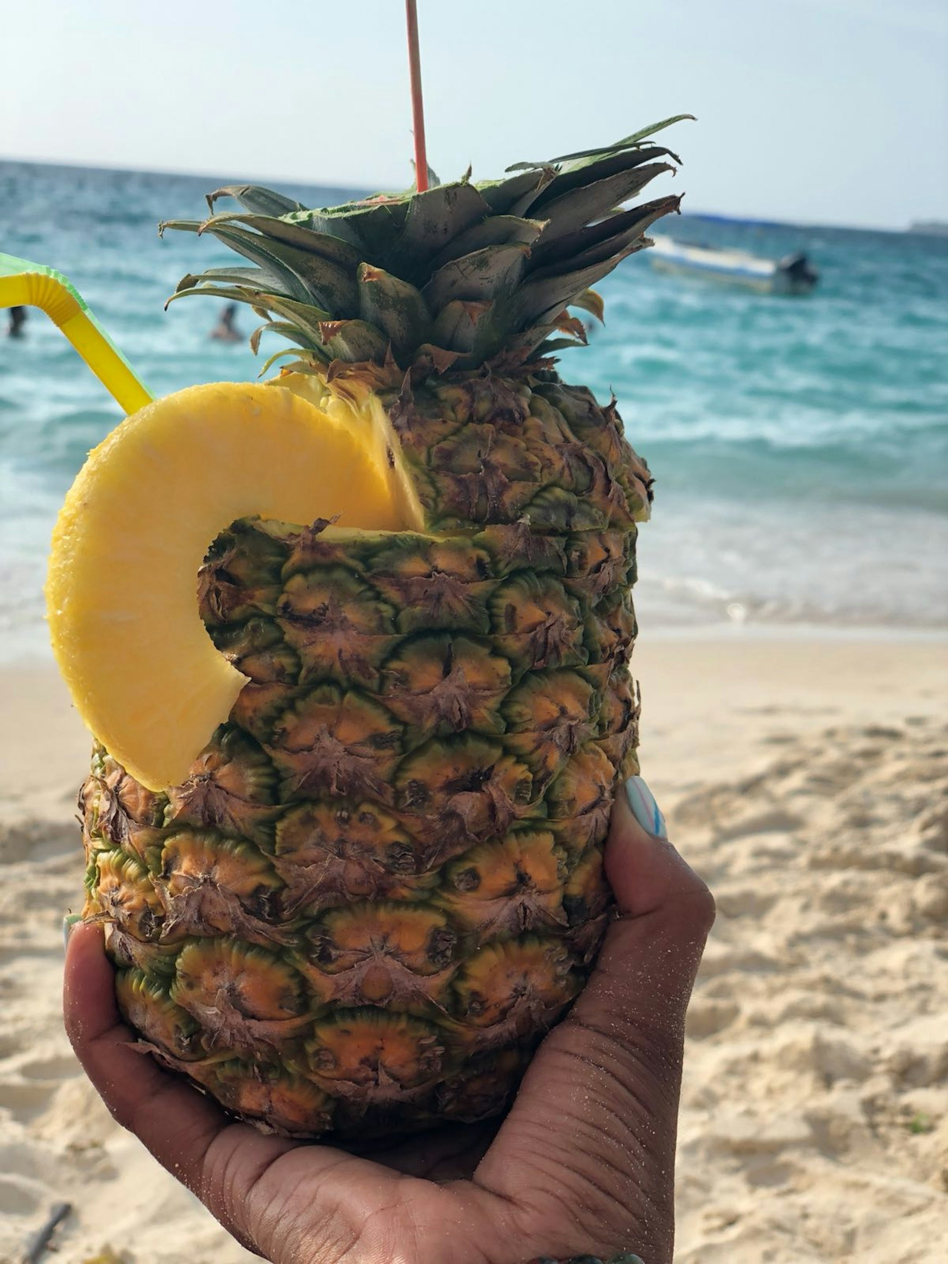 A hand holding a cocktail inside a pineapple, with a beach in the background – a necessity for a weekend in Cartagena