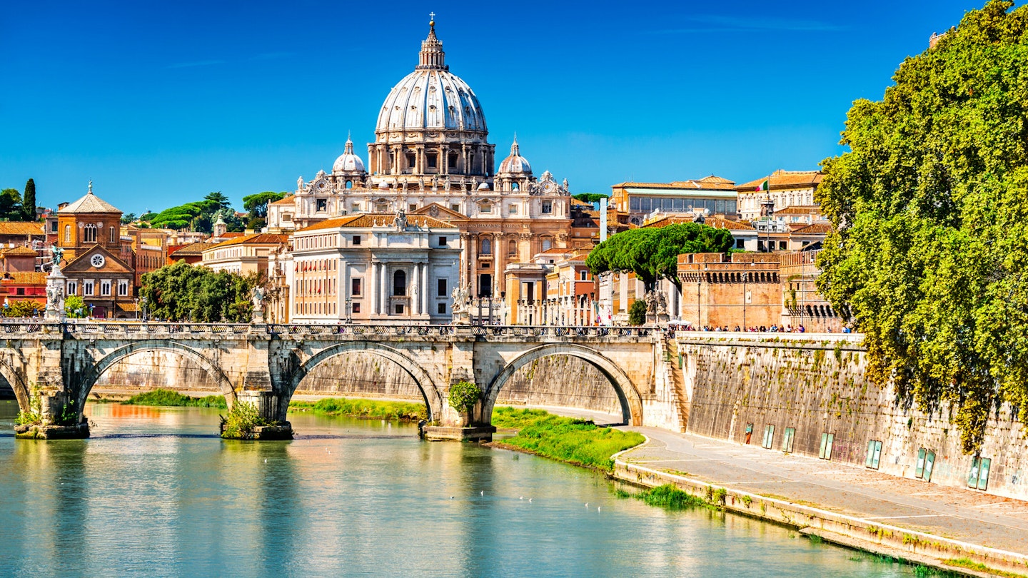 Under a brilliant blue sky the bridge of Pont Sant'Angelo crosses the calm blue waters of Tiber River from left to right, with St Peter's Basilica as a backdrop