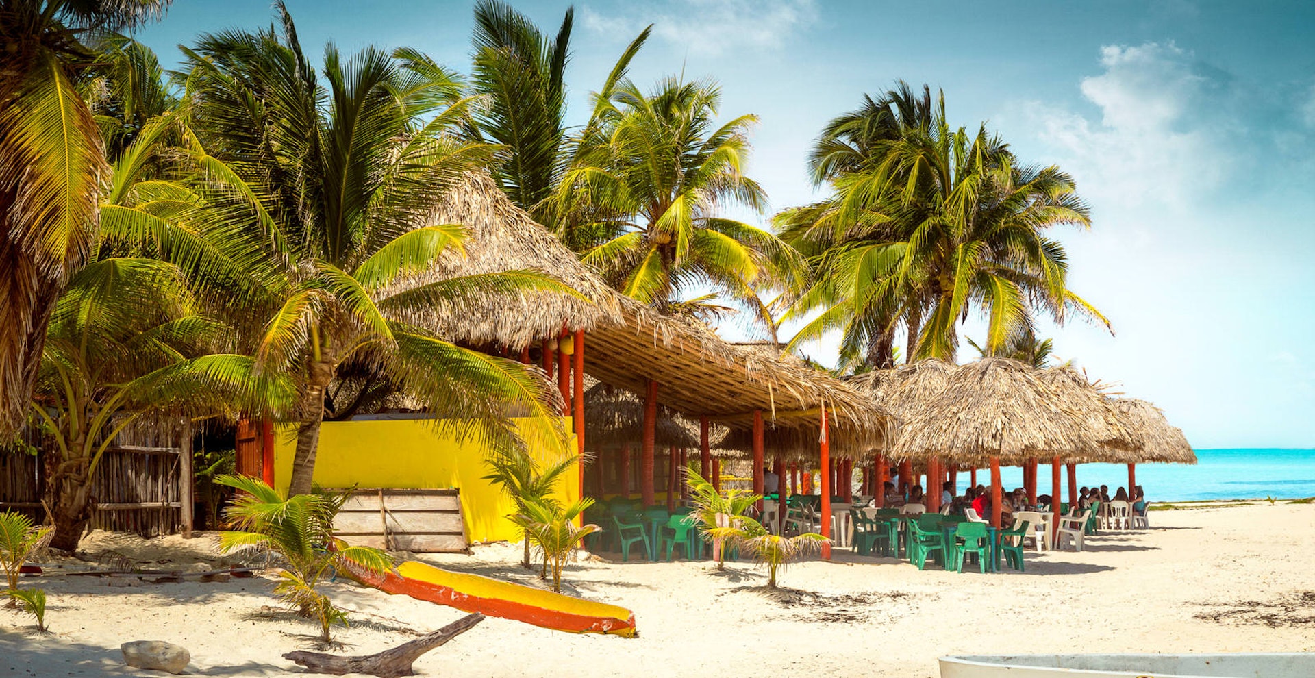 A thatched-roofed tropical bar sits beneath hulking palms on a beach at Cozumel island. The bar is painted bright yellow and red with green and white plastic chairs. It's partly obscured by green palm trees and sits on a white-sand beach with a jewel-blue sea in the background. 
