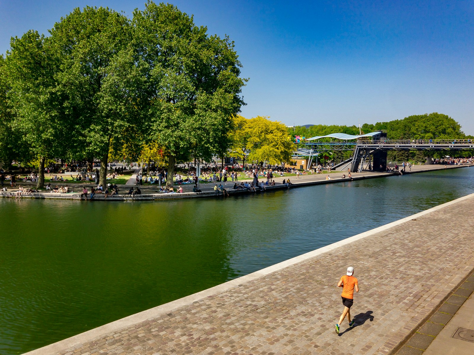 A lone man runs along the cobbled path flanking the Canal de l'Ourcq in Paris; the opposite side is covered in green trees 