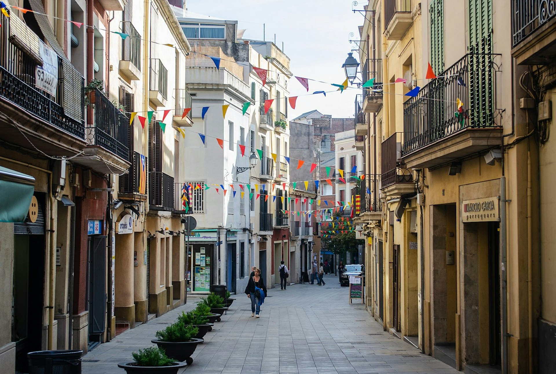 Looking down a narrow street, with shops on either side in elegant balconied buildings, and colourful bunting suspended across the street.