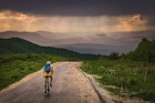 A man cycling alone in the mountains of Serbia on a rainy day; there are silhouetted peaks up ahead and greenery on either side of the road.