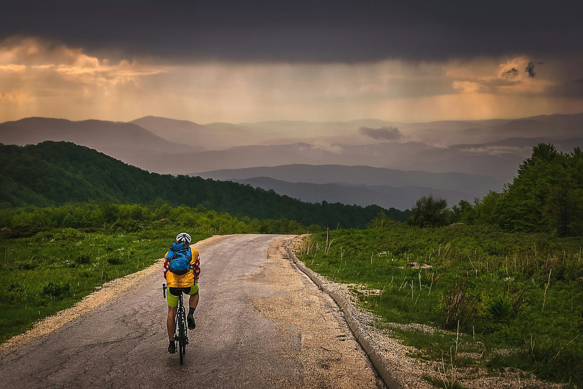 A man cycling alone in the mountains of Serbia on a rainy day; there are silhouetted peaks up ahead and greenery on either side of the road. EuroVelo