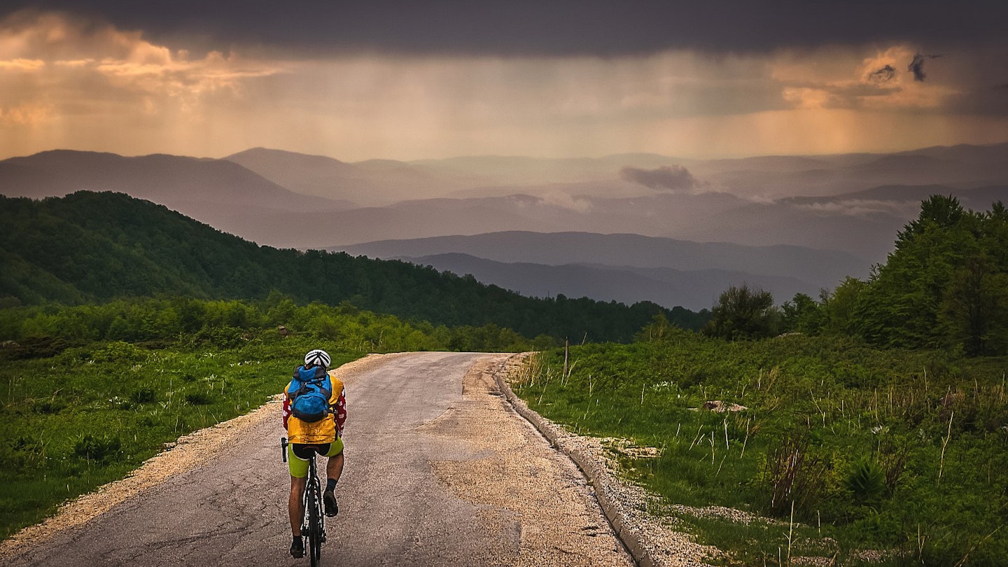 A man cycling alone in the mountains of Serbia on a rainy day; there are silhouetted peaks up ahead and greenery on either side of the road.
