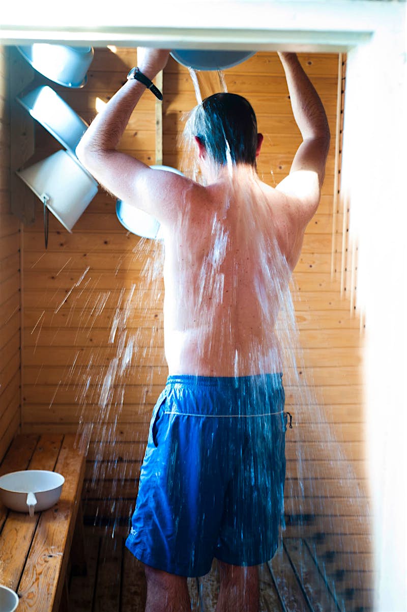 Features - showers-in-a-heatwave-2a06c65f0621 A man pours water over himself after a sauna. He is in blue swimming shorts and standing in a wood cabin.