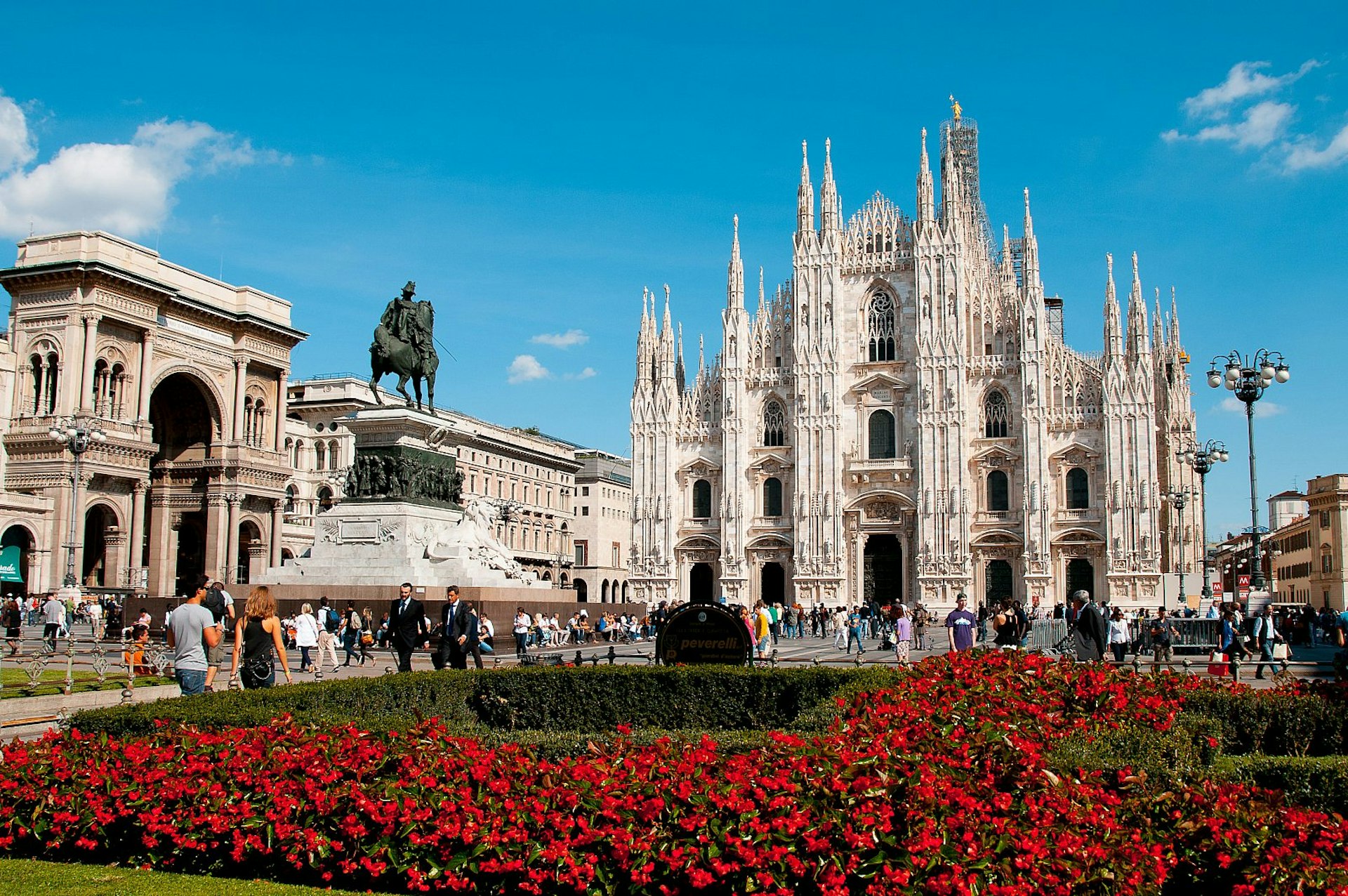 A red-flowering hedge in front of Milan's Duomo, a Gothic cathedral with a pearly white facade, extravagantly adorned with many spires and statues; there is a statue of a man on a horse to the side, and a large arch to the left.