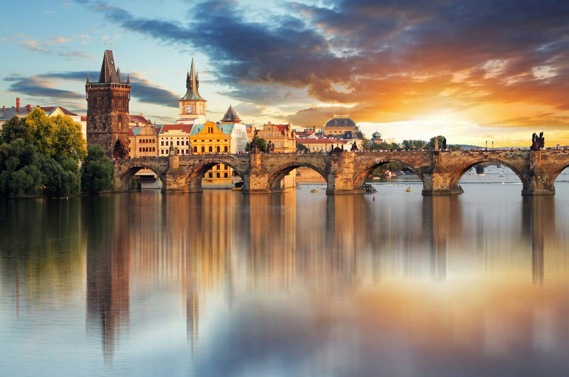 A panoramic shot along the river towards Charles Bridge, with the sun setting in a brilliant flash in the background; a dark cloud looms just above the sun