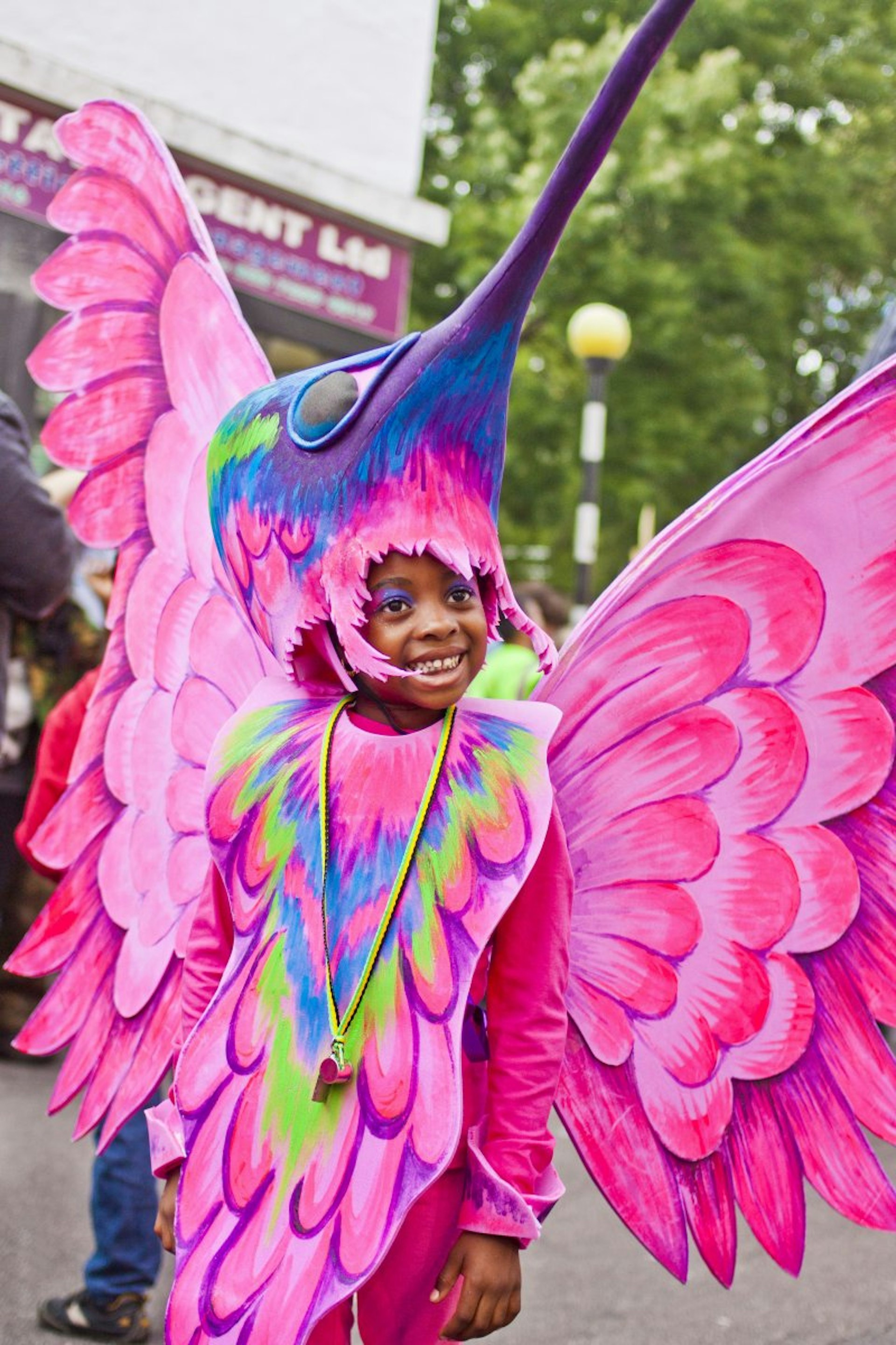 A child is dressed in a bright pink bird costume, with elaborate wings and a headdress including eyes and a blue beak