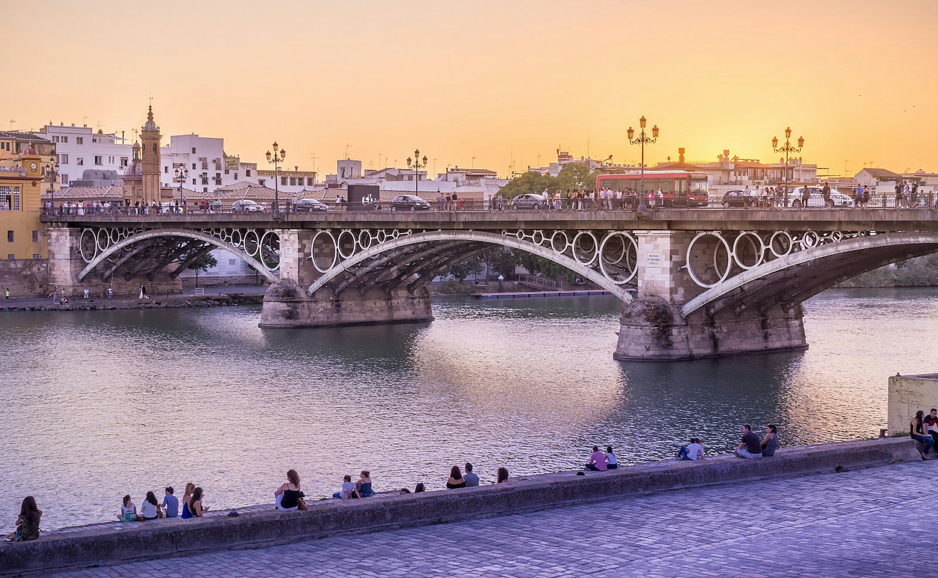 Sunset over the stone Triana bridge and the river Guadalquivir in Seville; in the foreground, people are sitting on a stone wall beside the river.