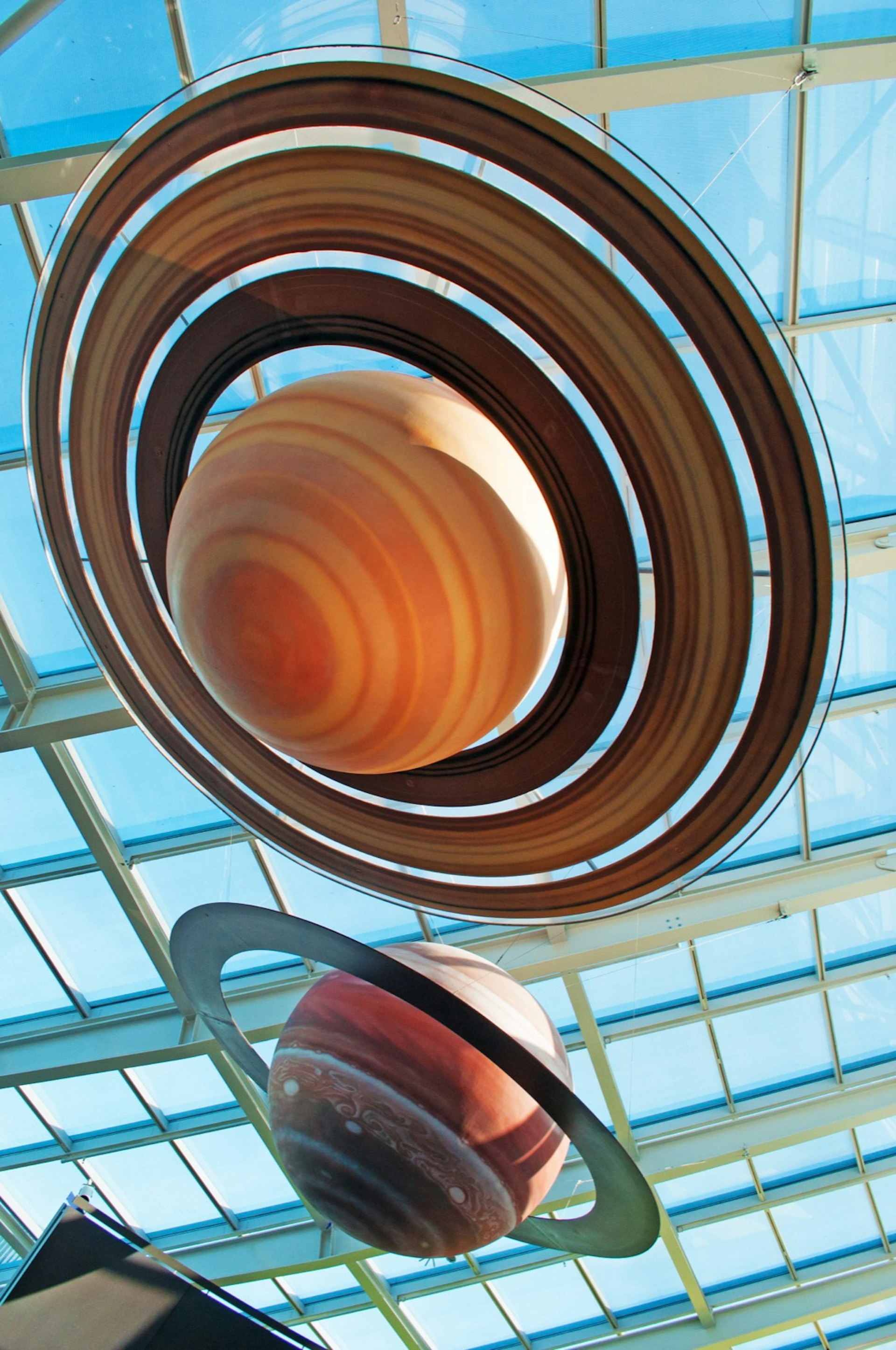 Reconstruction of the solar system at Adler Planetarium. The photos shows two ringed planets, one is orange and yellow with brown rings and the other is red and white with a green ring. The planets are suspended beneath a glass roof through which we can see a clear blue sky; Apollo anniversary experiences