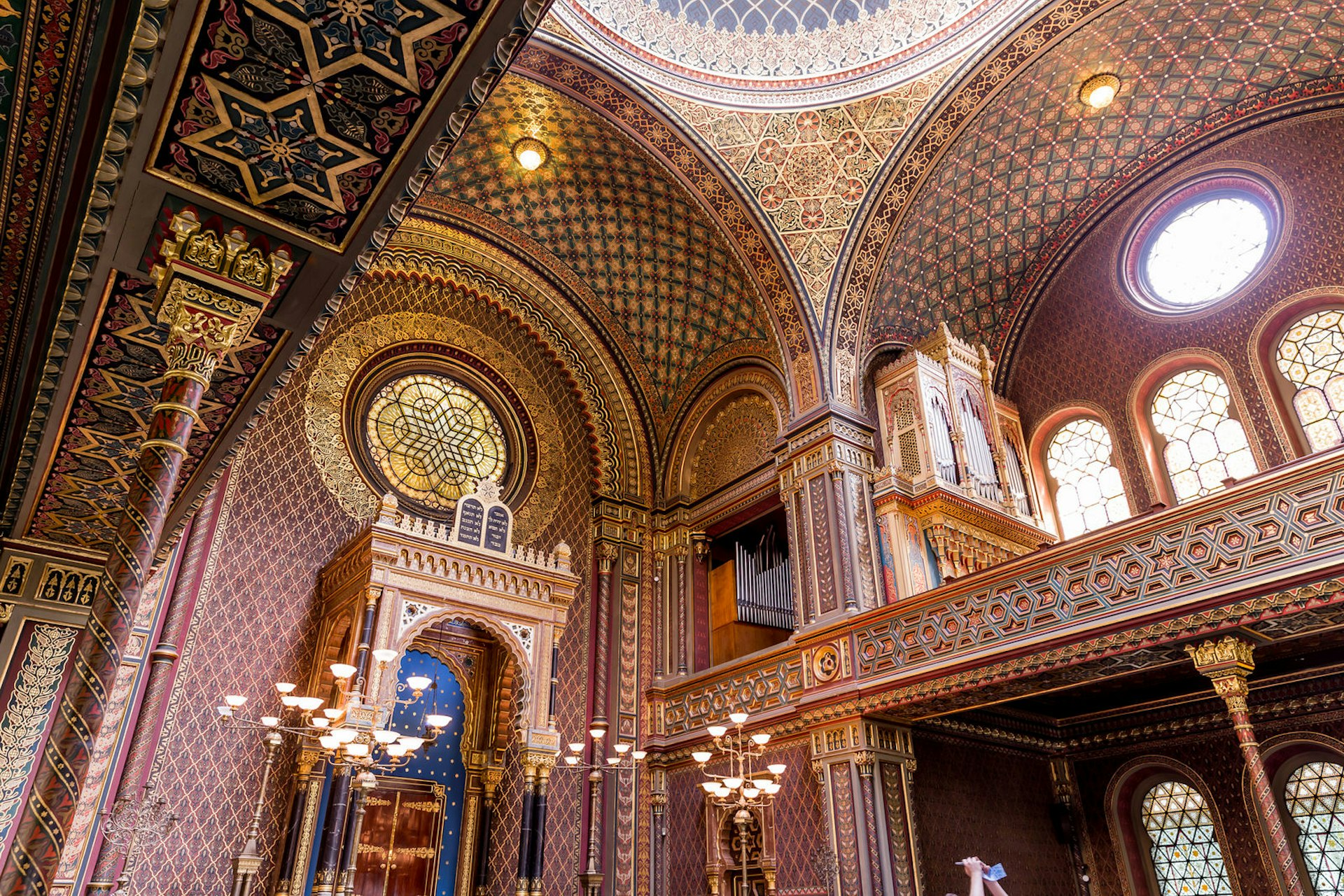 An incredible ornate interior of the Spanish Synagogue, with rich red tones mixed with elaborate gilded elements, the Star of David being a prominent feature; a large circular dome sits high above rounded stained-glass windows