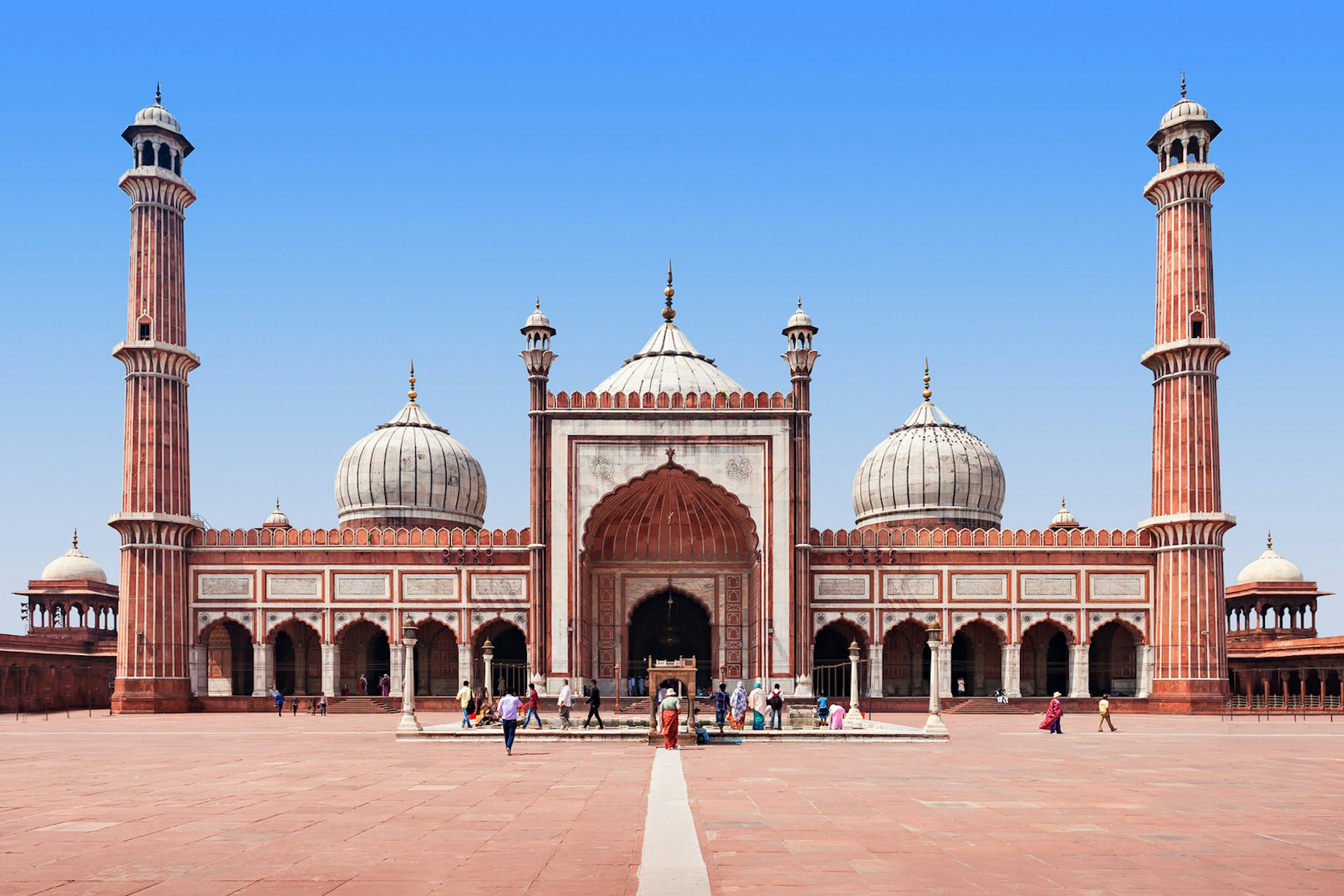 A large mosque in an open red-brick square. It has two tall minarets on either side and three domes
