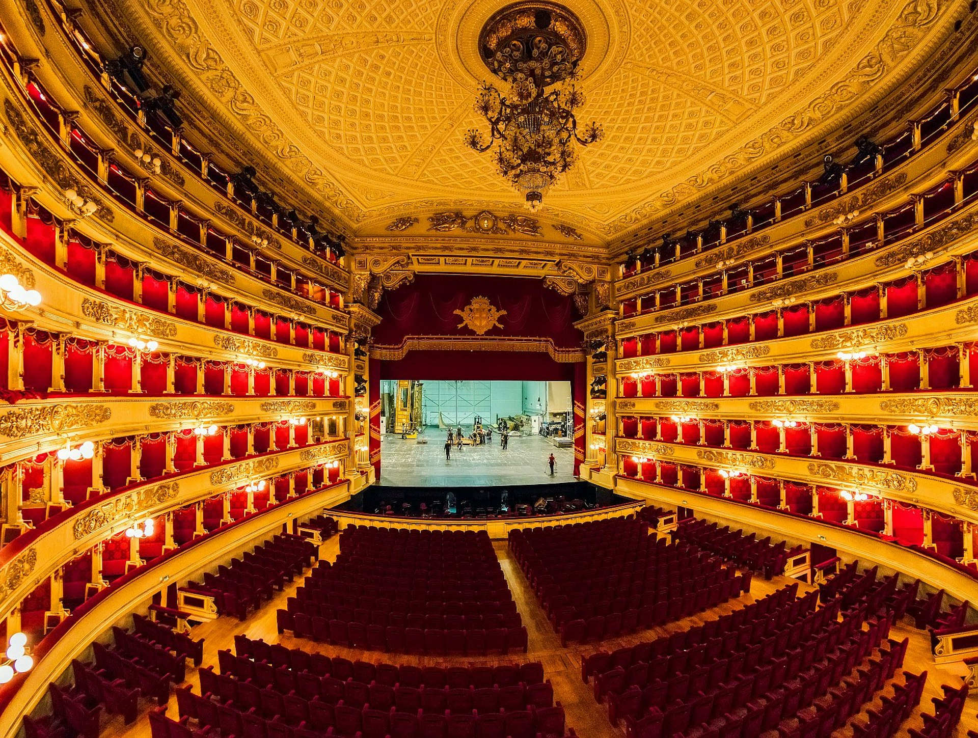 Looking out over the opulent interior of La Scala: a huge chandelier hangs from an ornate patterned ceiling; the walls are lined with six stories of red boxes; and there is read seating below. There are some workers on the stage.