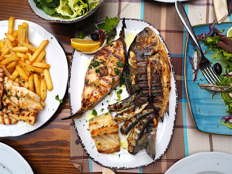 A plate of grilled fresh fish with a lemon garnish; another plate contains chicken and fries, while others contain green salads. They all rest on a wooden tabletop covered in a checked cloth.