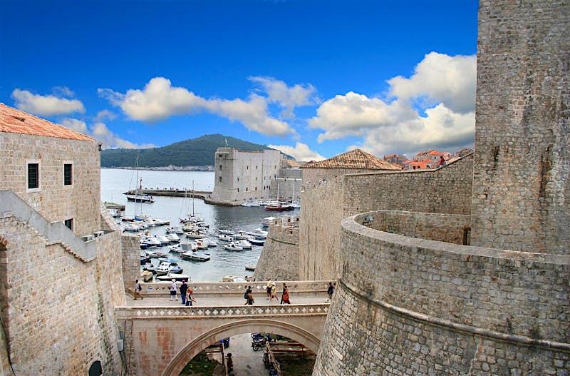 People walking across a stone bridge next to Dubrovnik's city walls; there is a harbour full of boats beyond with Lokrum island further still.