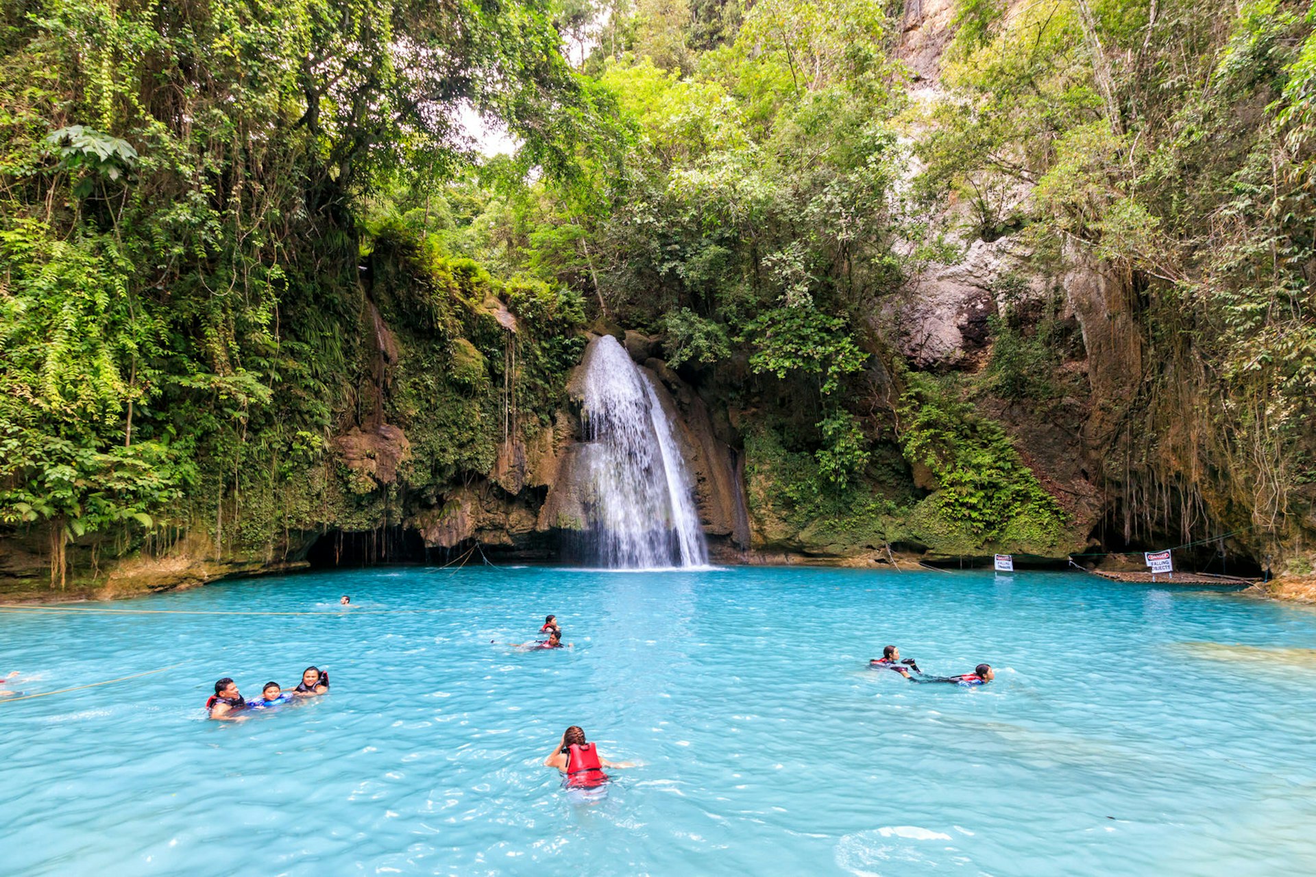 A shot of the blue pool at the bottom of Kawasan Falls, Cebu with a few people swimming in it. The blue waters are surrounded by lush greenery and we can also see the small waterfall. 