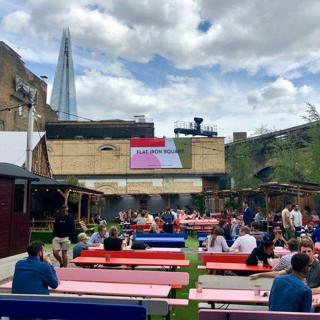 Several diners are sat at colourful benches in the outside area at Flat Iron Square. The Shard can be seen in the background. London's food halls and markets