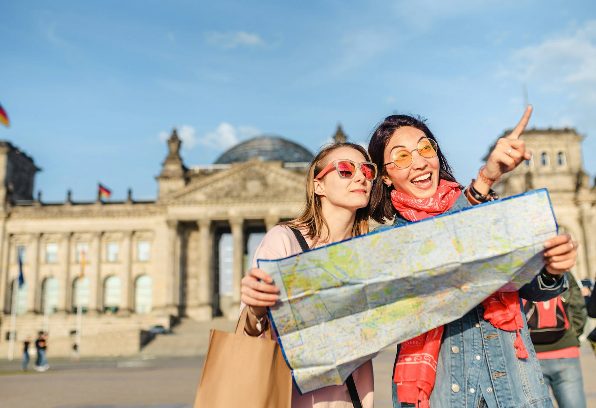 Two women wearing tinted sunglasses and holding a map with the Reichstag building in the background. One woman is pointing beyond the camera. It is a sunny day with a clear blue sky.