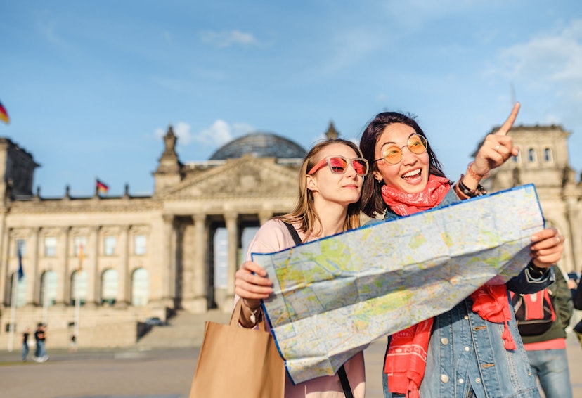 Two women wearing tinted sunglasses and holding a map with the Reichstag building on the background. One woman is pointing beyond the camera. It is a sunny day with a clear blue sky.
