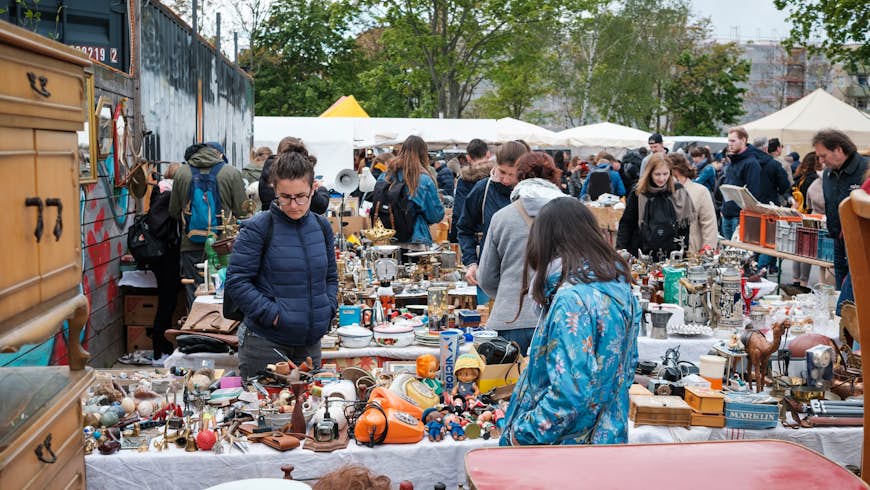 People browsing through item's at a flea market in Berlin's Mauerpark. People are wearing light jackets and looking at various items displayed on closely-packed tables. Some of the wares include; pots, toys, old-fashioned phones, ornaments and furniture.