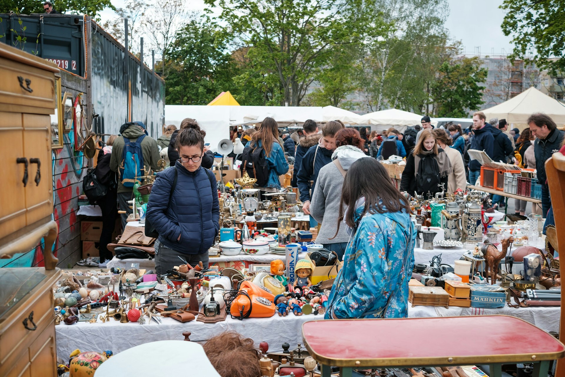 People browsing through item's at a flea market in Berlin's Mauerpark. People are wearing light jackets and looking at various items displayed on closely-packed tables. Some of the wares include; pots, toys, old-fashioned phones, ornaments and furniture.