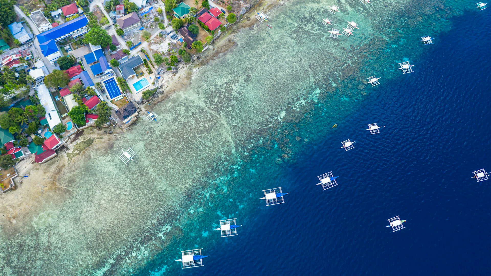 An aerial shot of Filipino boats floating on clear blue waters along the coast of Moalboal, Cebu. There is a narrow beach and colourful houses on the land.