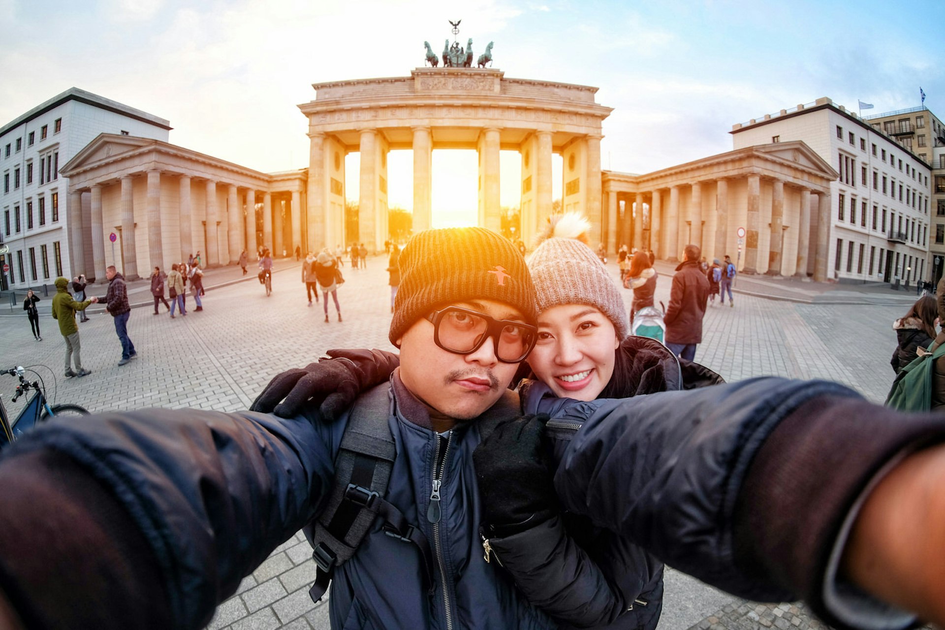 A couple taking a selfie in front of Brandenburg Gate, Berlin, at sunset. The man has his arms outstretched to hold the camera and the woman has her arms wrapped around him. They are both wearing warm coats and woolly hats.