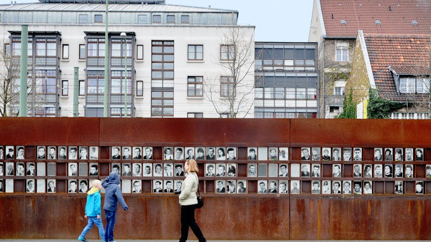 A photo of the Berlin Wall Memorial with three people walking by. The memorial is a metal wall contructed where the Berlin Wall once stood showing black and white photos of people. 