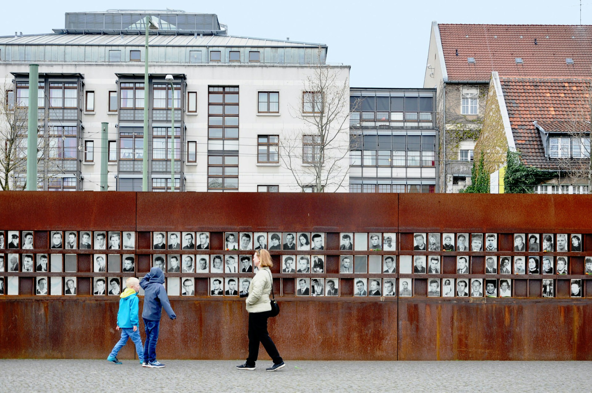 A photo of the Berlin Wall Memorial with three people walking by. The memorial is a metal wall contructed where the Berlin Wall once stood showing black and white photos of people. 