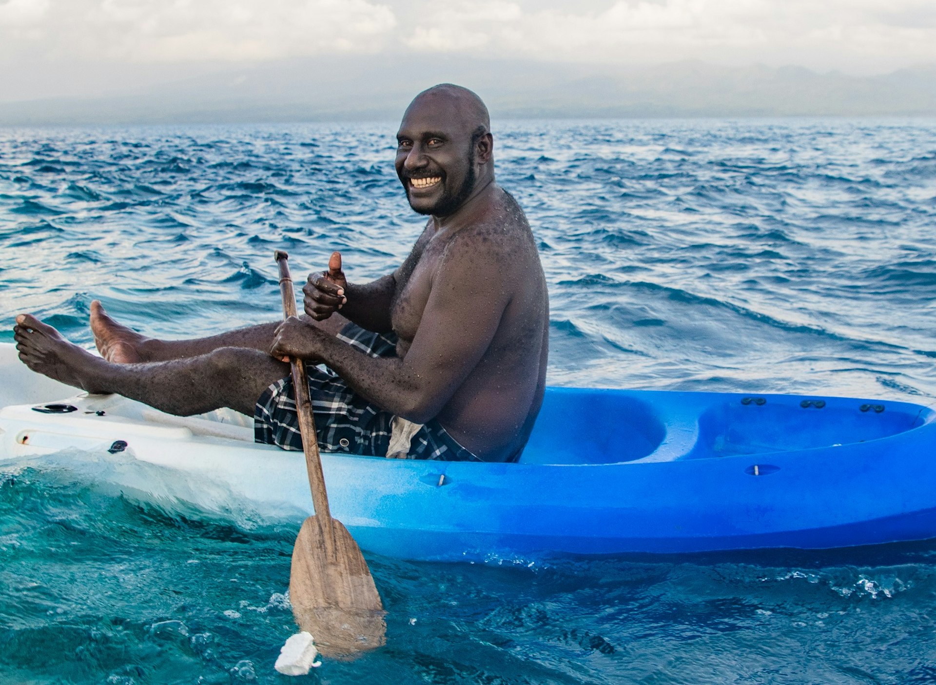 A man in a plastic kayak that fades from blue to white, holding a wooden paddle, grinning at the camera. The man is surrounded by the Pacific Ocean and there are white clouds on the horizon