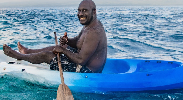 A man in a plastic kayak that fades from blue to white, holding a wooden paddle, grinning at the camera. The man is surrounded by the Pacific Ocean and there are white clouds on the horizon