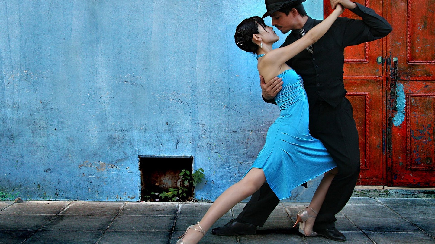 Tango dancers in the La Boca area of Buenos Aires; the man is dressed in black trousers, shirt and hat with a black and white spotted tie; the lady wears a blue dress which matches the blue wall behind them.
