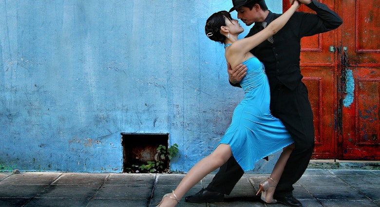 Tango dancers in the La Boca area of Buenos Aires; the man is dressed in black trousers, shirt and hat with a black and white spotted tie; the lady wears a blue dress which matches the blue wall behind them.