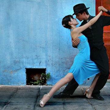 Tango dancers in the La Boca area of Buenos Aires; the man is dressed in black trousers, shirt and hat with a black and white spotted tie; the lady wears a blue dress which matches the blue wall behind them.