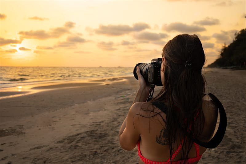 A young woman points an SLR camera towards a sunset over a tropical beach